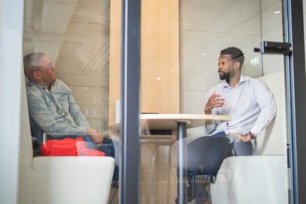 Twee mannen zitten in een glazen vergaderruimte aan de Joop Geesinkweg en voeren een gesprek.